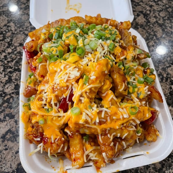 Waffle fries topped with orange sauce, shredded cheese, and green onions in a white takeout container.
