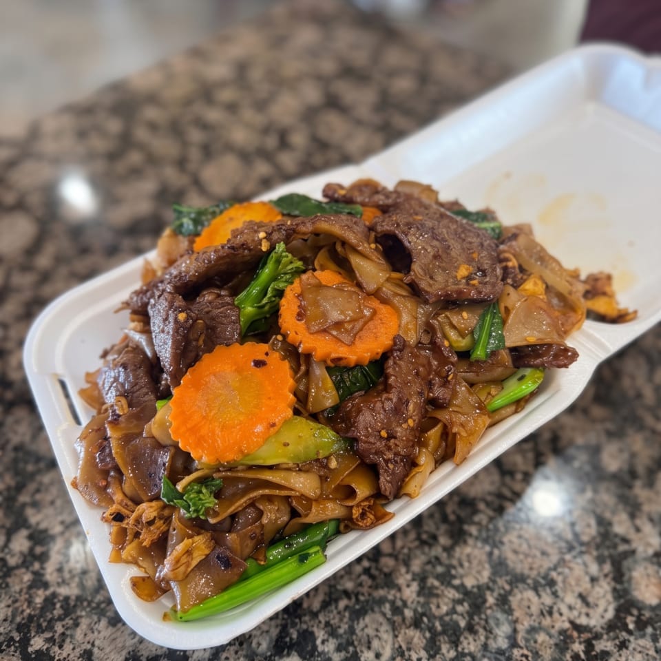 Beef Pad See Ew with wide noodles, carrots, and Chinese broccoli in a white takeout container.