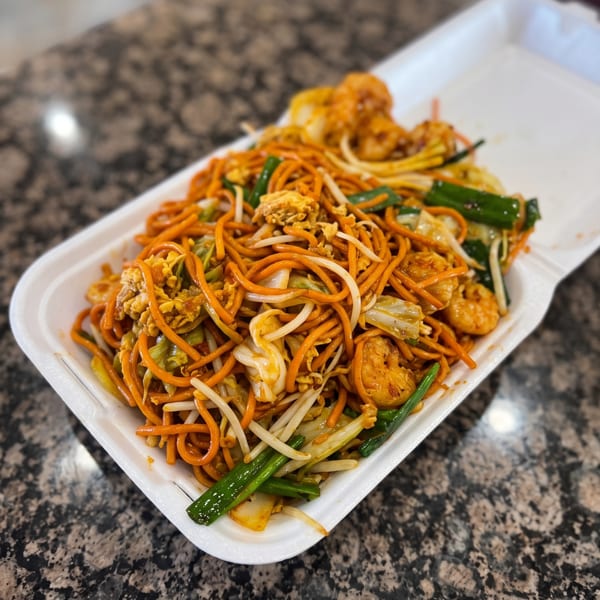Shrimp lo mein with vegetables and egg in a white takeout container on a speckled counter.